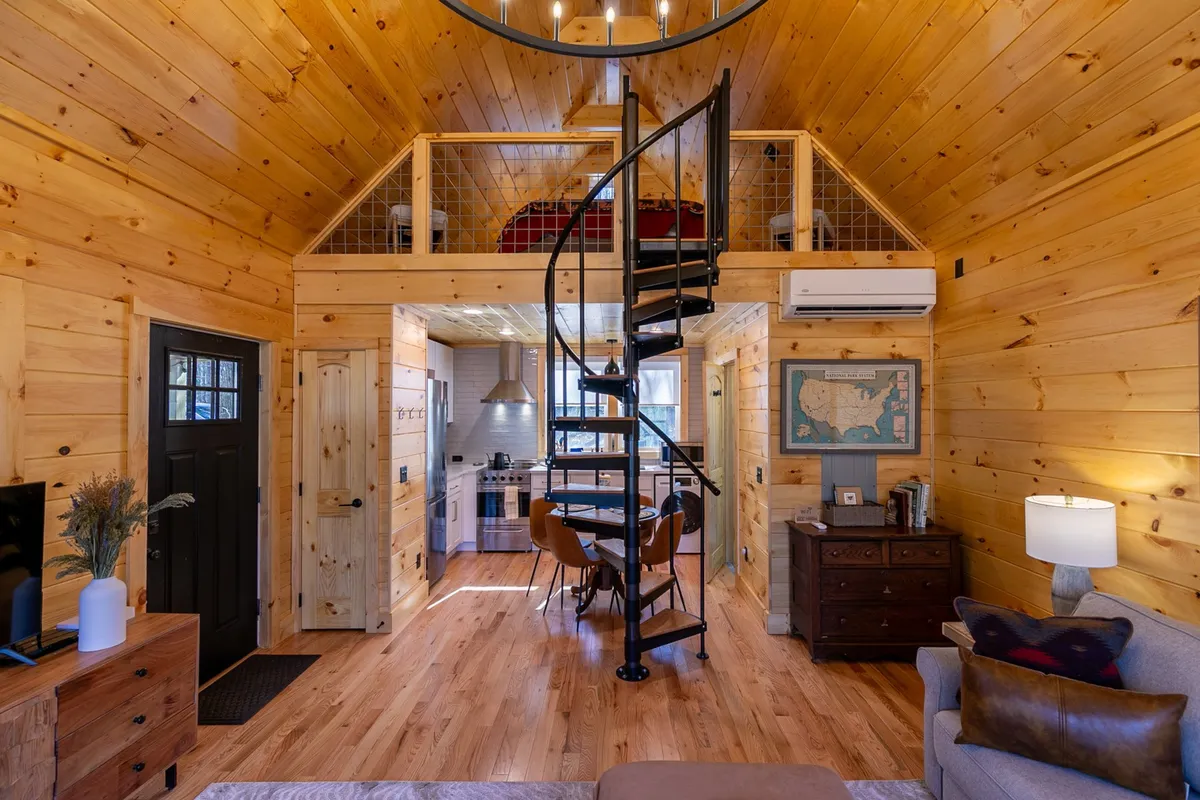 A welcoming open-concept space featuring a spiral staircase, a dining area with a wooden table, wood-paneled walls, and a view into the integrated kitchen. The seating and table arrangement foster social interaction, while the natural wood tones and light create a warm ambiance.