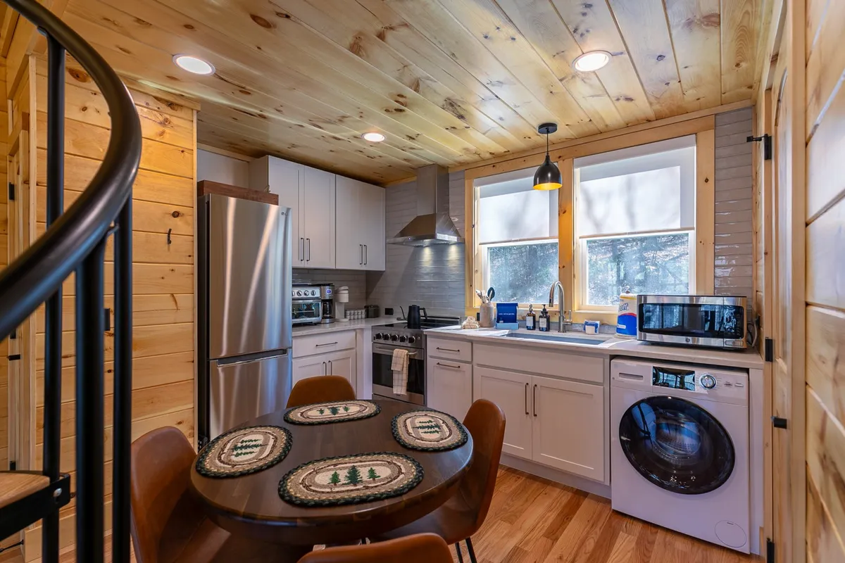 This interior shot features a round wooden dining table, stainless steel kitchen appliances, and large windows that flood the area with sunlight. The windows provide glimpses of nature, reinforcing the indoor-outdoor connection. The setting is both functional and inviting, with space for cooking and socializing.