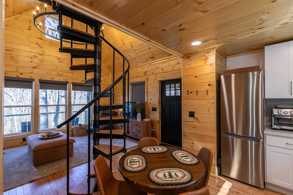 A wide-angle view of the spiral staircase leading up to the loft, flanked by a dining table, large windows, and warm wooden walls. The staircase becomes a focal point, inviting guests to ascend to the sanctuary above while also showcasing the open design and the visual connection between spaces.
