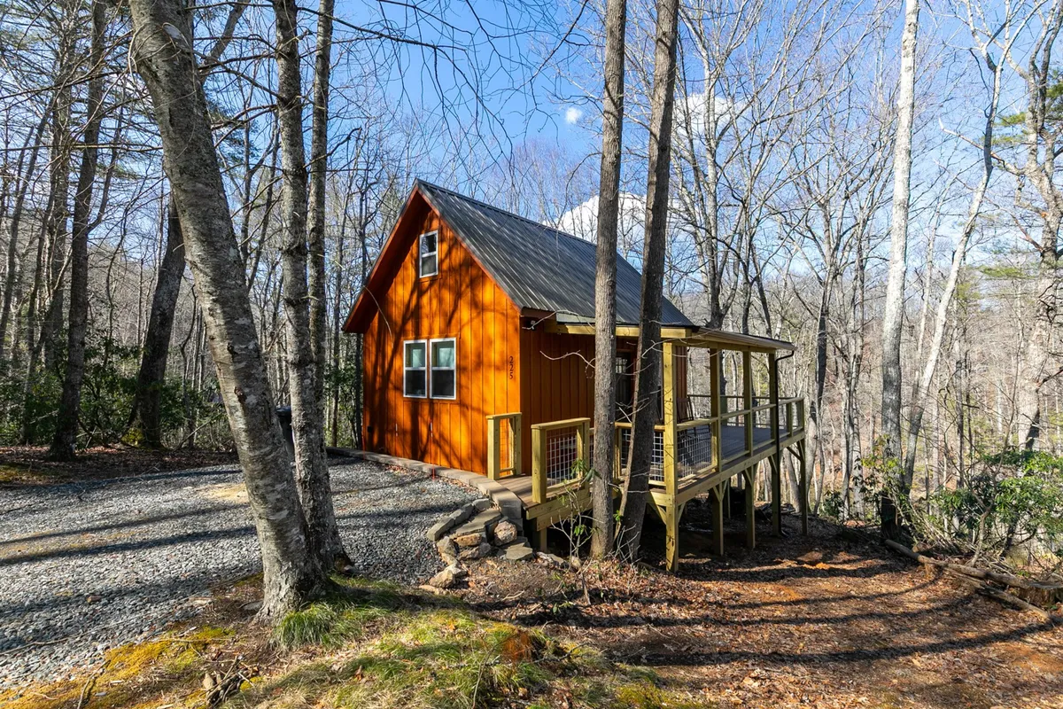 A bright orange cabin stands out among a backdrop of lush green trees, its spacious deck area offering a welcoming space for relaxation. The setting exudes a cozy charm, with the porch and yard hinting at opportunities to connect with the outdoors, enjoy morning coffee, or gather around an evening campfire. The natural surroundings emphasize a getaway vibe close to nature.