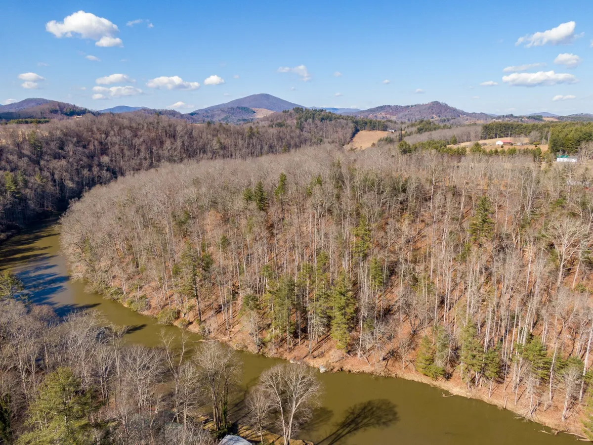 A tranquil view of the New River winding through lush greenery, with a tree line and distant mountains beneath an open sky. The scene captures the serene beauty of the area and provides a glimpse of the natural landscape visitors can explore nearby.