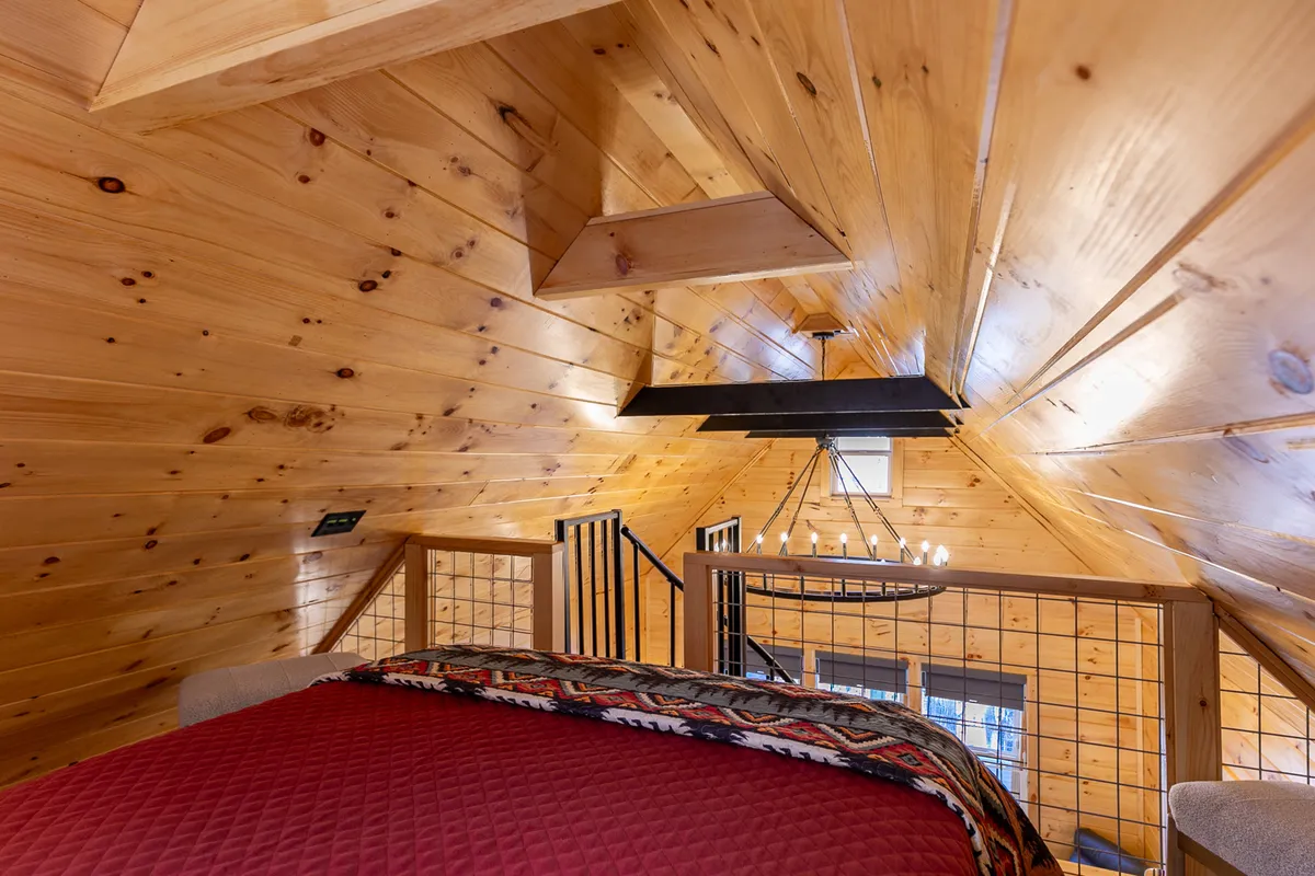 A cozy loft bedroom with a wooden ceiling, a queen bed adorned with a rich red quilted bedspread, and a delicate chandelier. The bed is positioned to enjoy treetop views through windows, allowing natural light to fill the space. The rustic yet romantic decor creates an intimate, inviting atmosphere that feels tucked away yet open to the rest of the cabin.