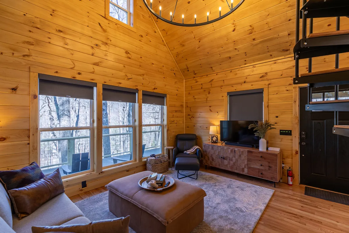 This image highlights a spacious living room with wooden walls, large windows that let in ample sunlight, a comfortable sofa, a television, and a chandelier. The expansive windows frame the outdoors, filling the space with natural light and offering scenic views. The seating area is arranged for relaxation and social gatherings.