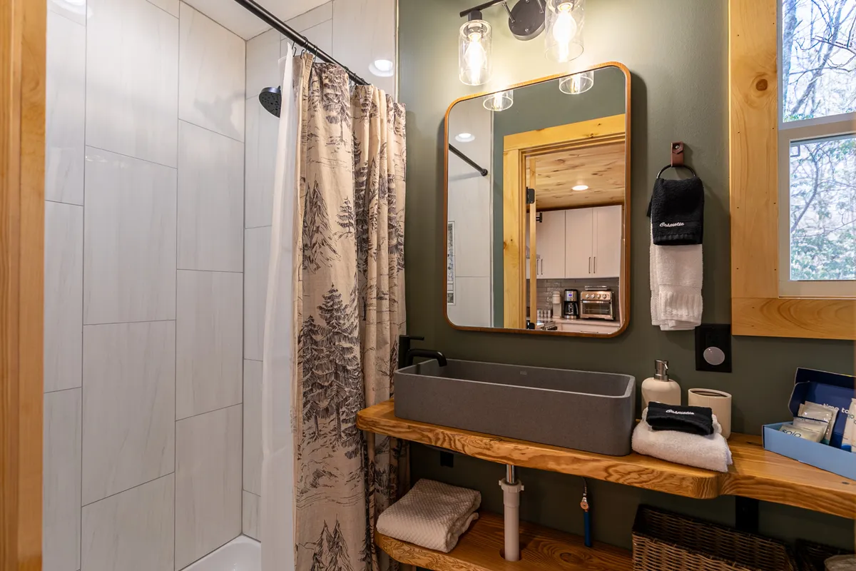 This image displays a calm and serene main-level bathroom with a shower, large mirror, and a wooden sink stand. The space feels inviting and clean, with thoughtful details that support relaxation and self-care routines. Elements such as plush towels and a tidy setup contribute to the sense of guest comfort and convenience.