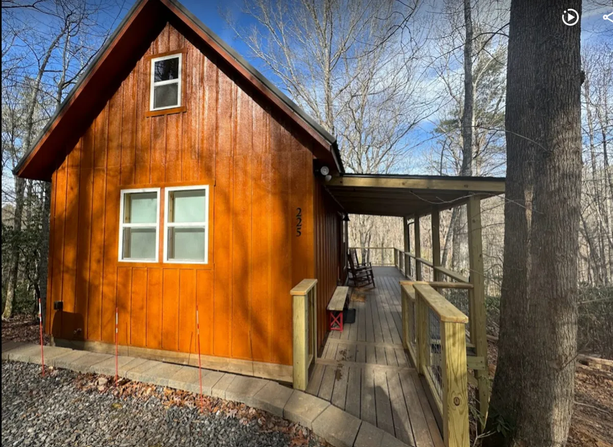 A rustic wooden cabin with a charming front porch, surrounded by tall trees. The inviting porch hints at activities like morning coffee, sunset watching, or stargazing after a day outdoors.
