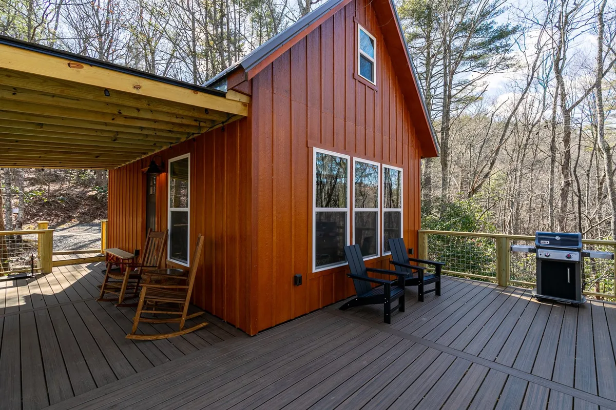 The exterior of the cabin with a prominent deck seating area, grill, and large windows. Trees surround the space, providing a secluded, tranquil feel for both morning coffee and sunset dining outdoors.