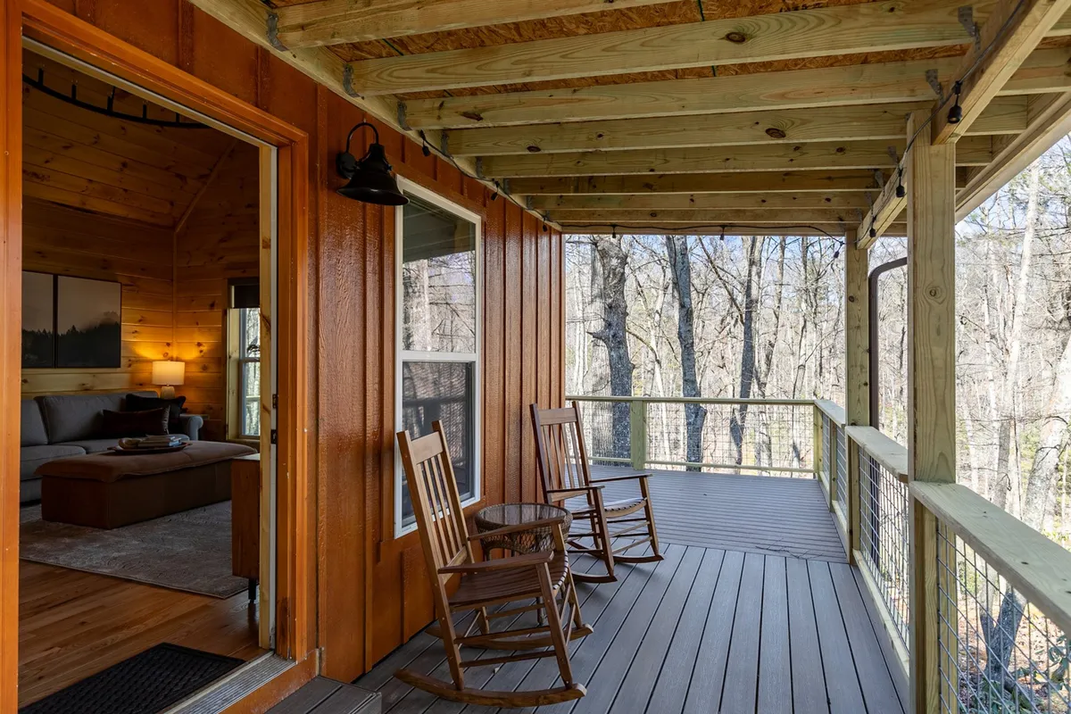 A peaceful deck scene with rocking chairs overlooking the treetops, perfect for relaxing with a morning coffee or sharing stories at dusk. The surrounding forest and partial interior glimpse create a sense of seamless indoor-outdoor living.